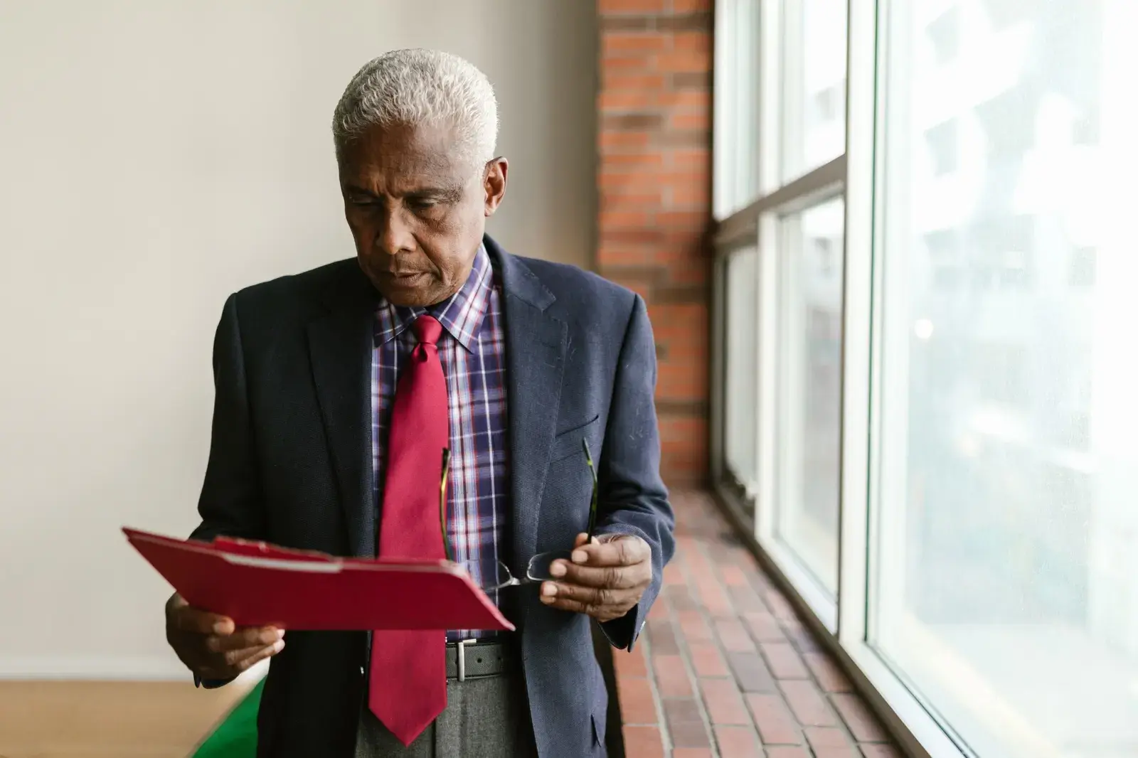 Man in a suit reading a document from a red folder beside a window